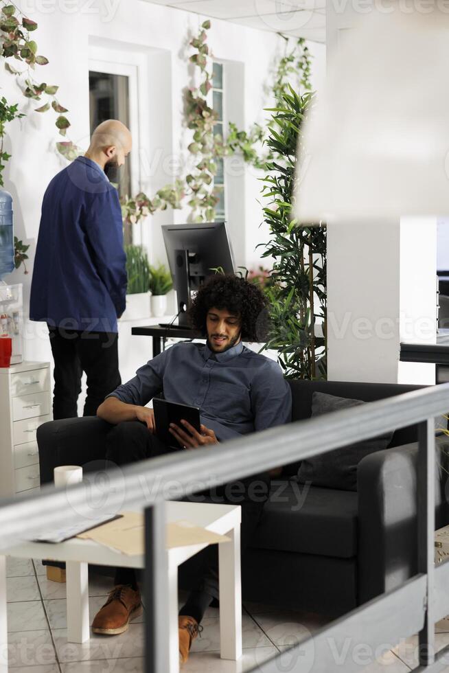 Arab start up company worker using digital tablet during work break while sitting on couch. Young entrepreneur chatting in social media network in business open space office photo