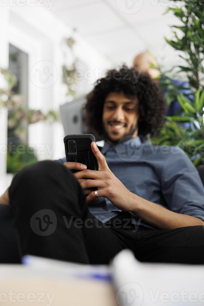 Smiling businessman relaxing in office, using smartphone to send message. Young arab executive manager texting in social media on mobile phone while having break in coworking space photo