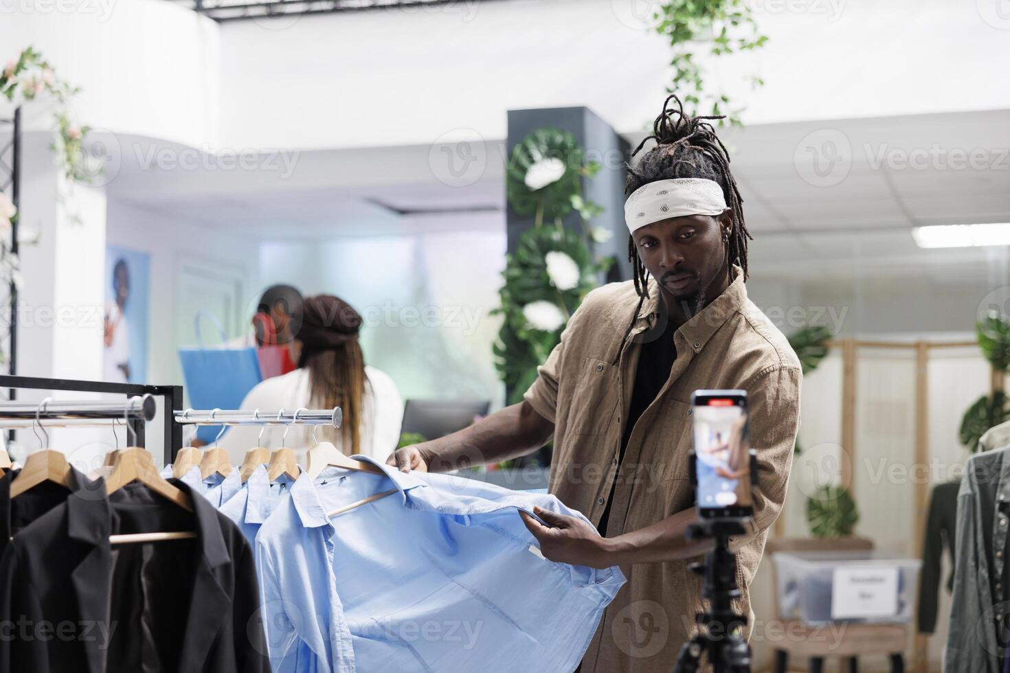 Social media blogger recording video with shirts review for brand promotion in shopping mall. African american marketing specialist speaking about apparel fabric quality on smartphone camera photo