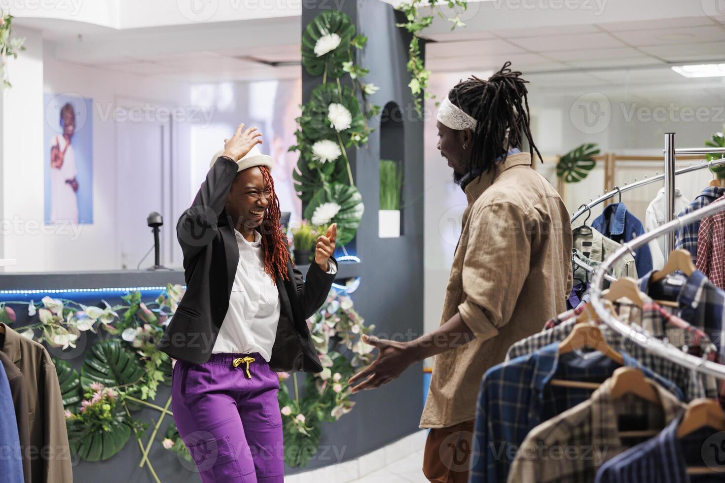 Cheerful woman laughing and wearing hat while shopping with boyfriend in clothing store. Smiling african american customer trying on accessory and asking friend for advice in mall photo