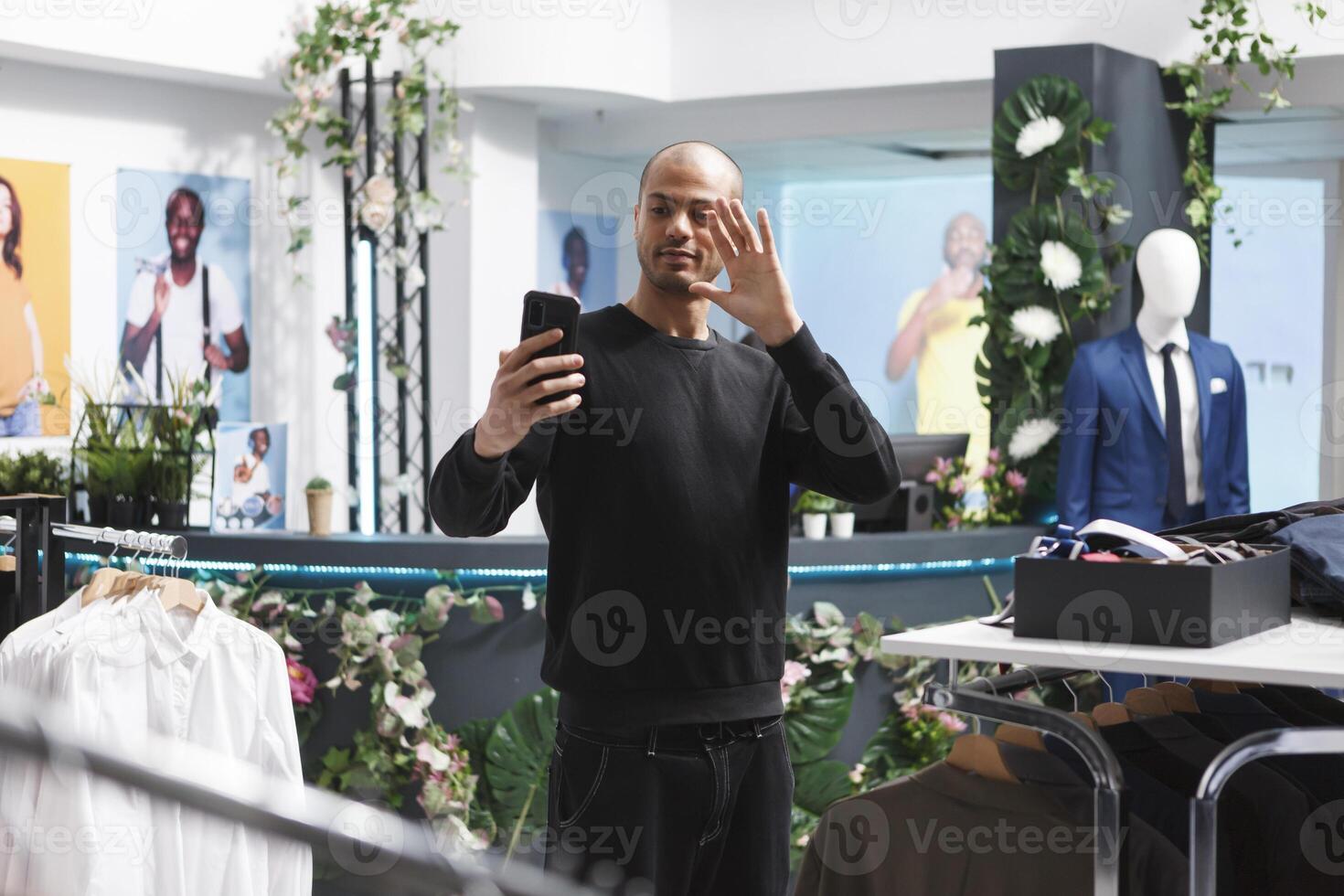 Arab man customer in clothing store holding smartphone, looking at front camera and waving with hand while shopping for apparel. Influencer greeting followers while streaming in boutique photo