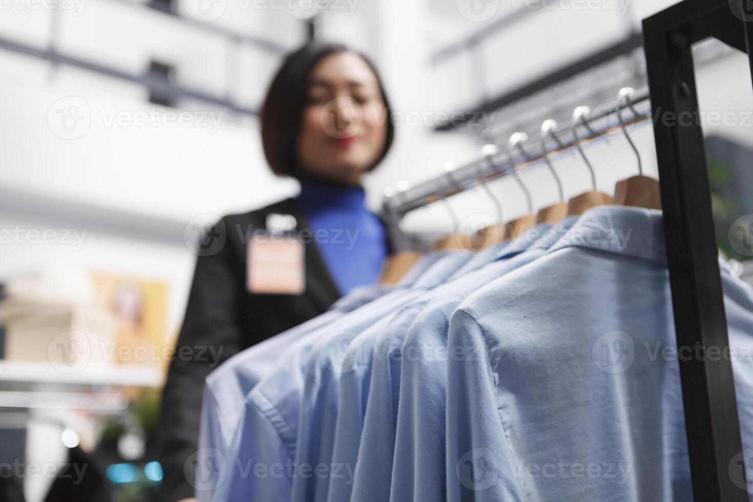 Formal shirts hanging on display rack while asian woman assistant working in clothing store selective focus. Menswear garment on hangers in shopping mall boutique closeup photo