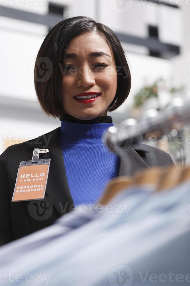 Fashion boutique smiling asian woman seller with badge examining formal shirts on display rack. Department shopping center clothing store assistant managing garment inventory photo