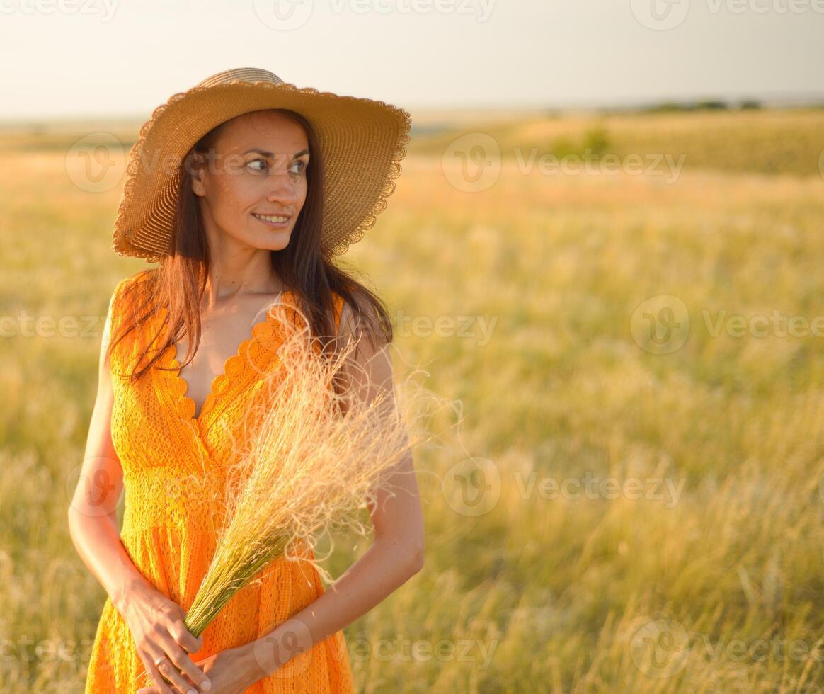 Young woman in an orange dress and a straw hat standing on a field in the rays of the setting sun photo