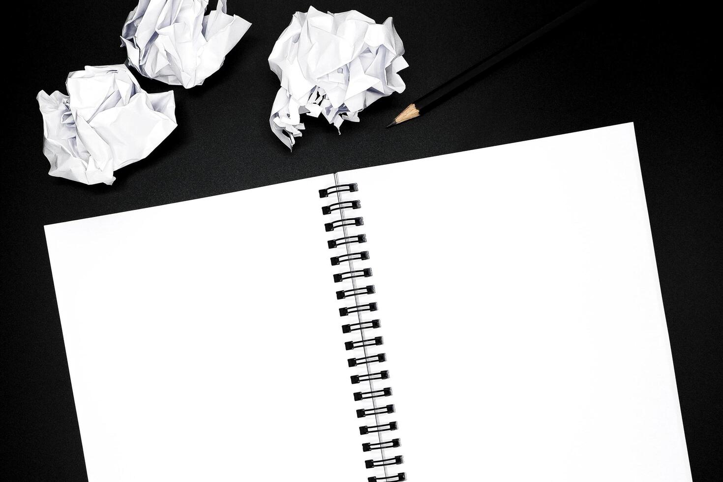 Blank spiral notebook with black pencil and crumpled papers on black background. photo