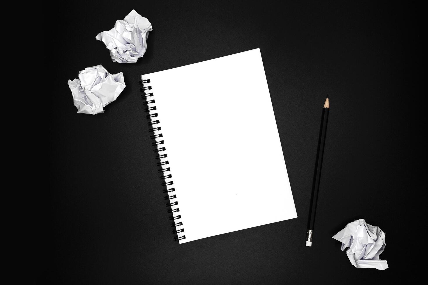 Blank spiral notebook with black pencil and crumpled papers on black background. photo
