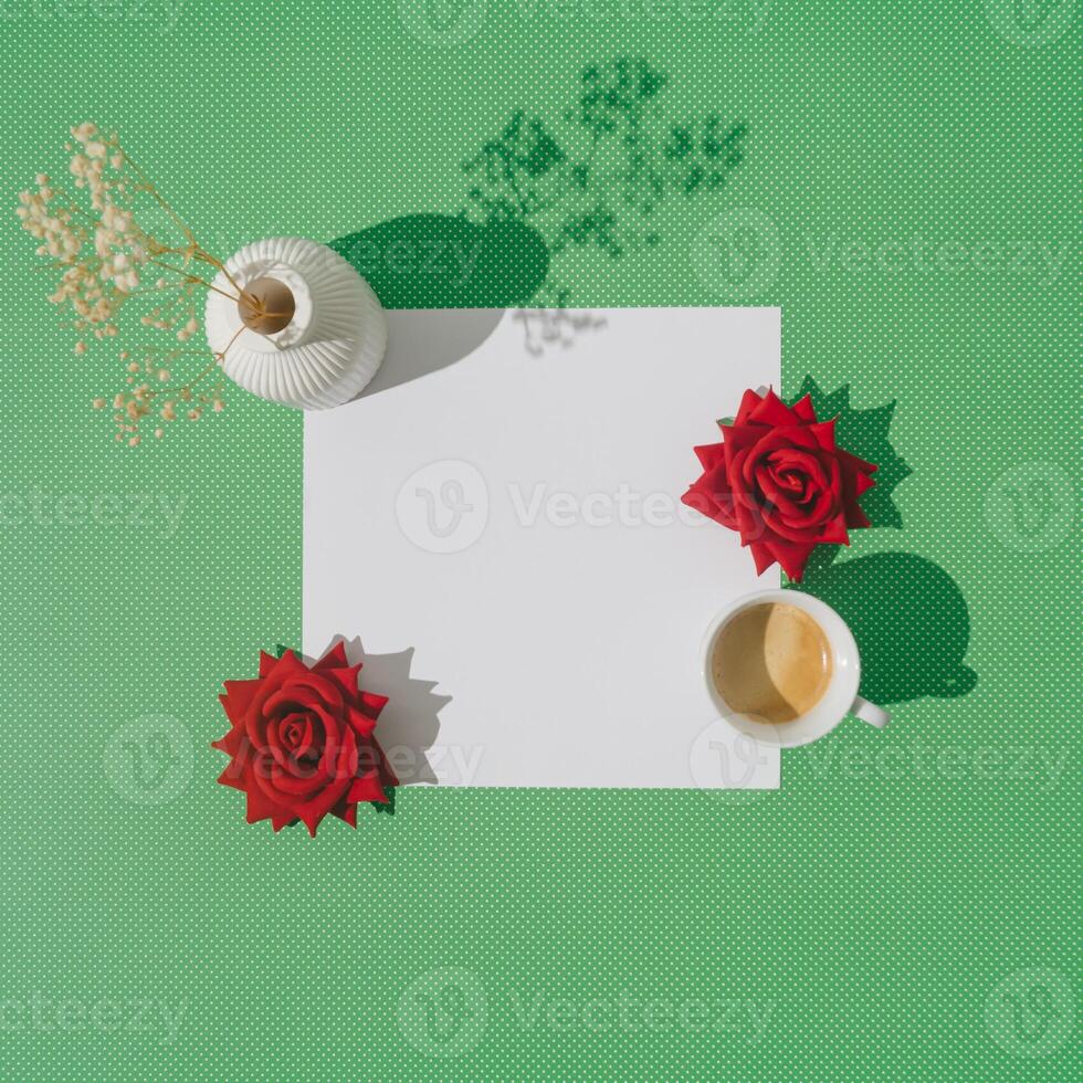 Creative spring layout made with cup of coffee, red roses, white vase with dry flowers and paper card note copy space on green background. Minimal concept. Trendy coffee with flowers idea. Flat lay. photo