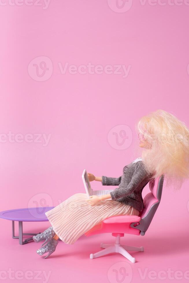 Doll with laptop in chair on pink background. Working at home, freelance creative concept. photo