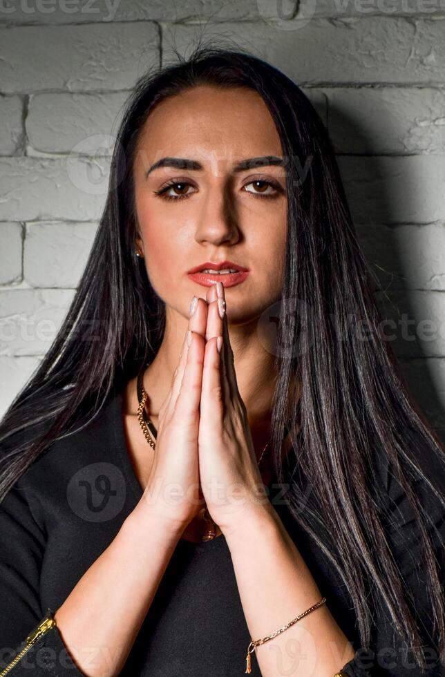 Girl brunette in black dress near the white brick wall folded her hands in prayer photo