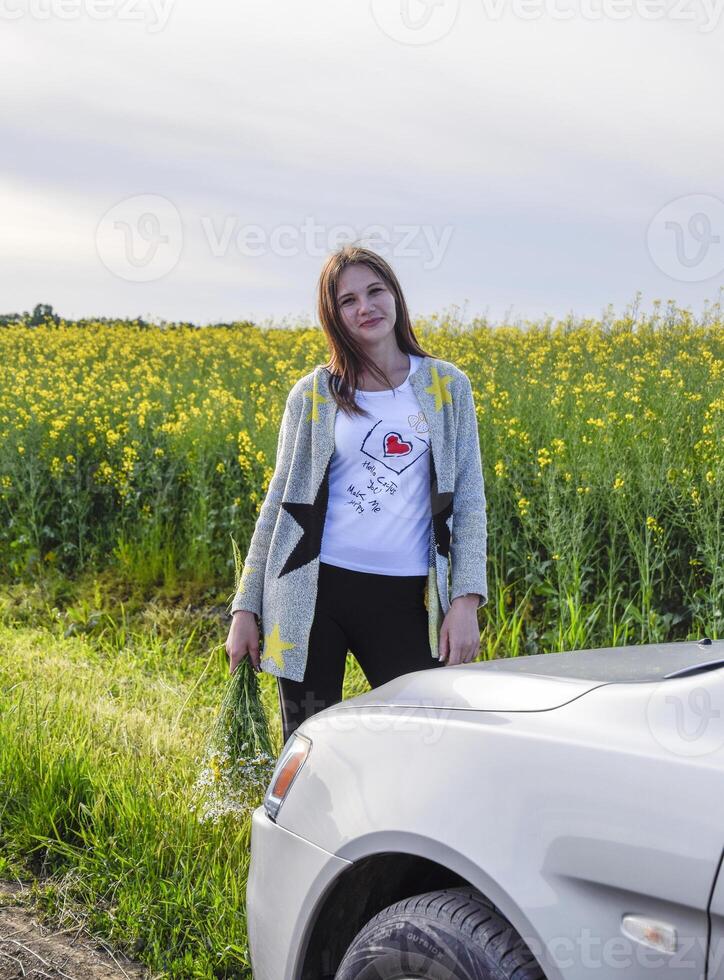 A beautiful young woman with a bouquet of daisies stands near a silver car photo