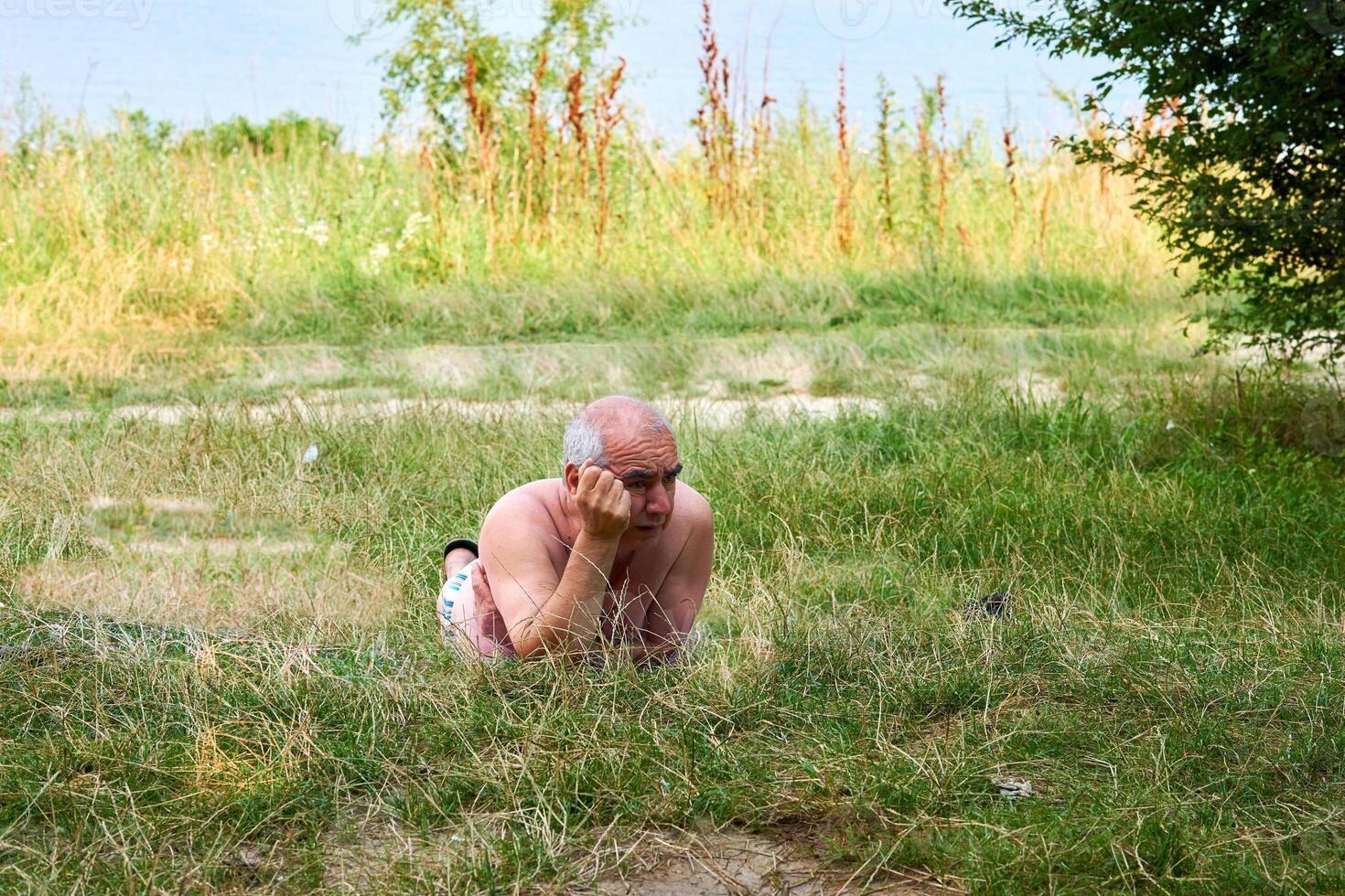 un mayor hombre es descansando en un césped en un césped cerca un verano río foto