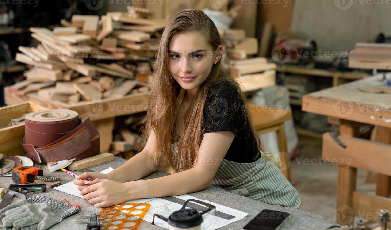 A young woman is training to be a carpenter in the workshop. She works with a laptop computer in a wood workshop. female carpenter contact customers by smartphone. SME orders, Start-up and small photo