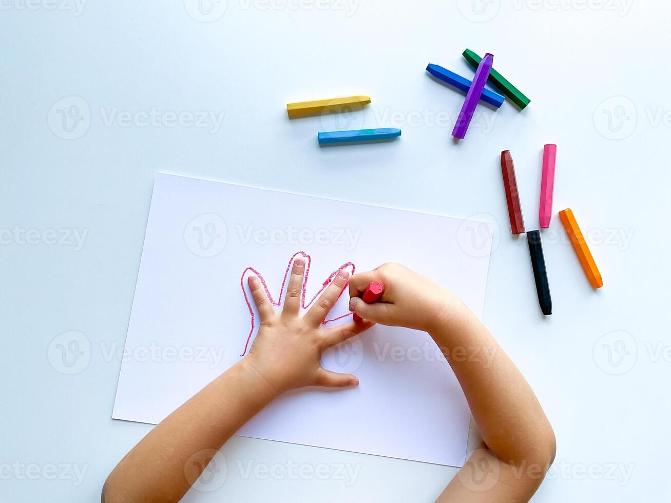 Childrens hands draw their hand with wax crayons on white paper, top view. photo