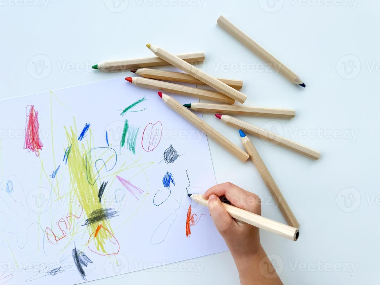 small child draws with colored pencils on paper on white table. photo