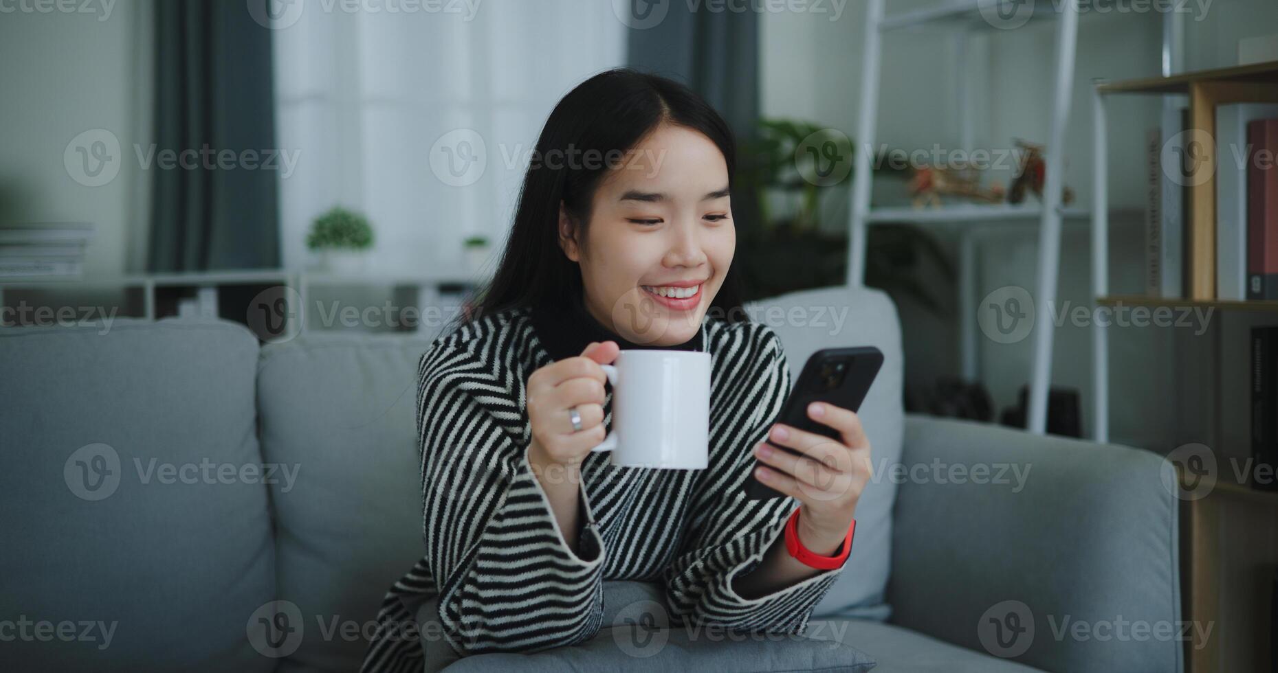 retrato de contento joven asiático mujer sentar en sofá disfrutar chateando en móvil teléfono mientras Bebiendo café en Mañana en vivo habitación, gratis tiempo, toma descanso, sonriendo foto