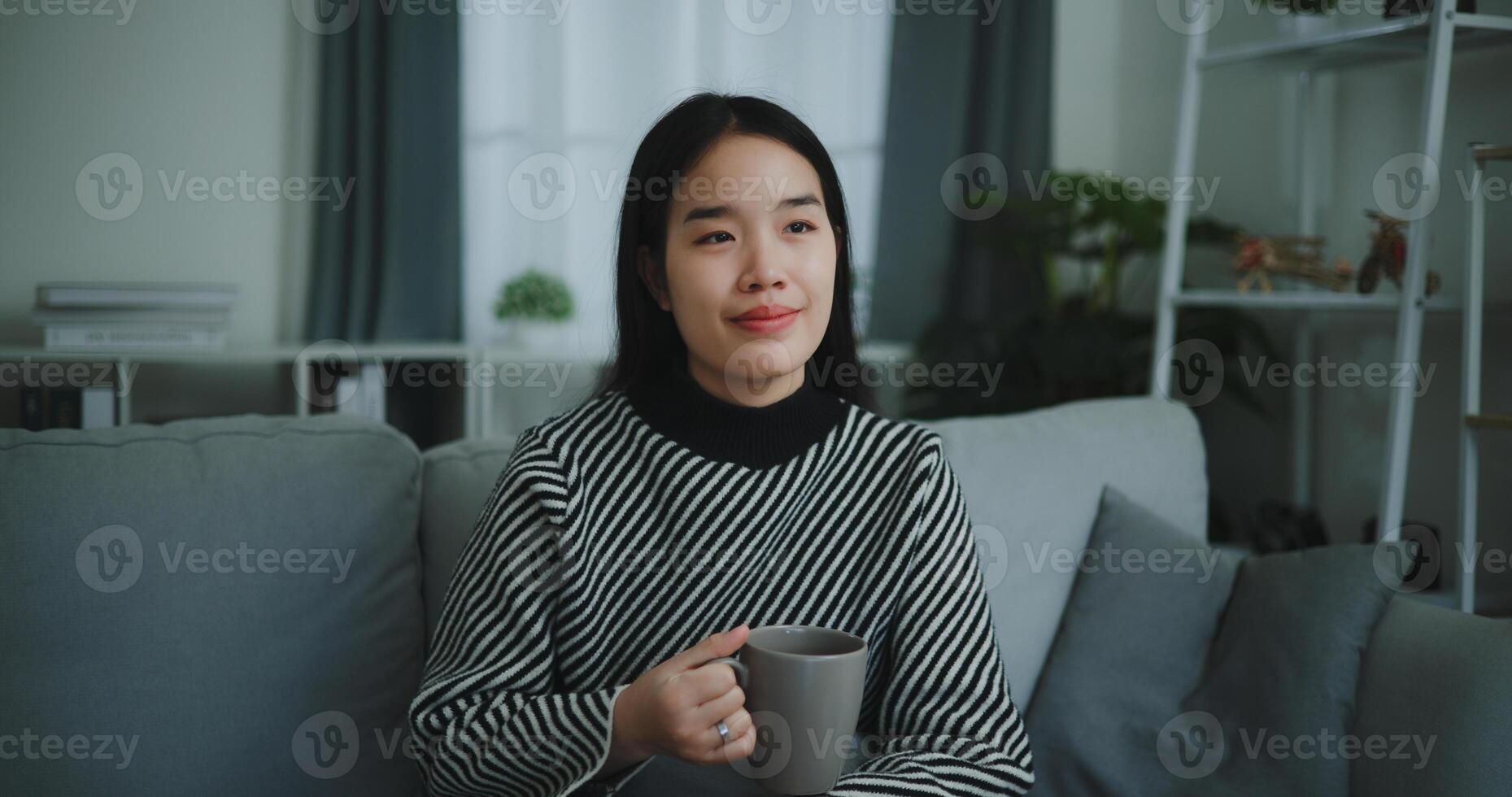 retrato de relajante joven mujer participación taza disfrutar oler café o té y Bebiendo con felicidad en Mañana mientras sentar en sofá en vivo habitación, gratis tiempo, toma descanso en casa, sonriendo foto