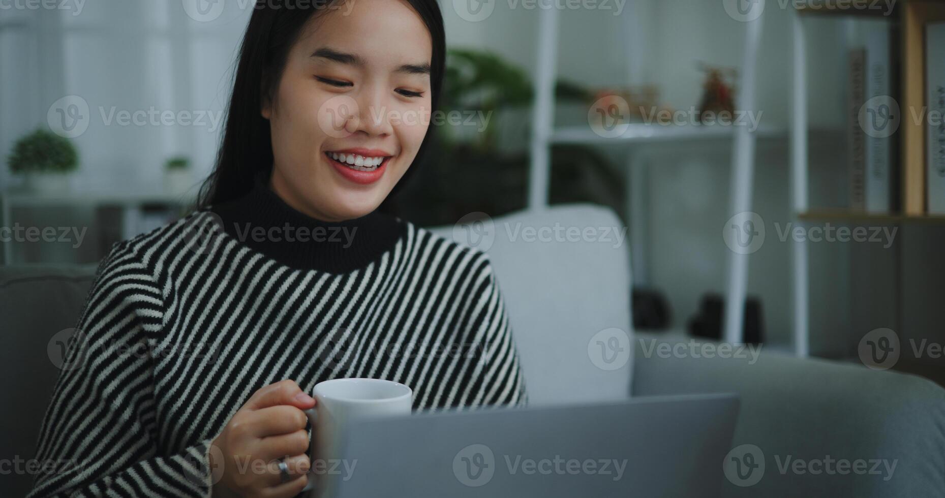 Portrait of Happy young asian woman sit on sofa drinking coffee or tea while working creative job from home on laptop with online network,take break,smiling photo