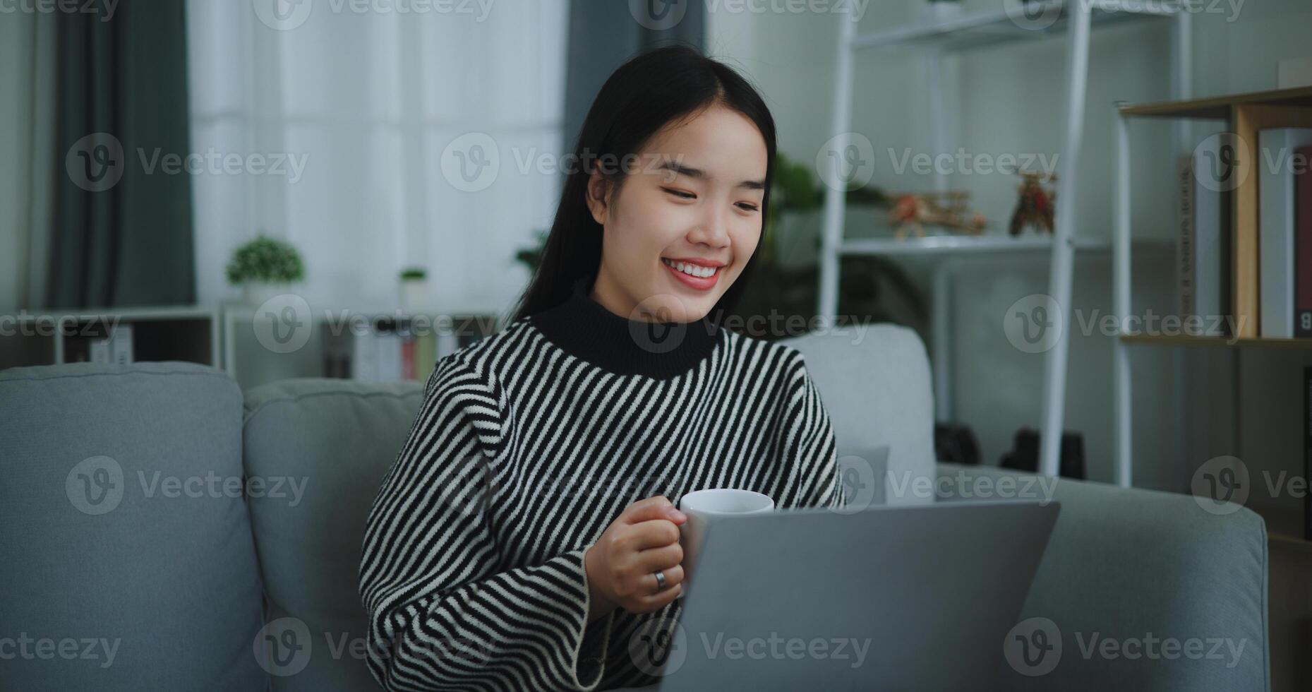 Portrait of Happy young asian woman sit on sofa drinking coffee or tea while working creative job from home on laptop with online network,take break,smiling photo
