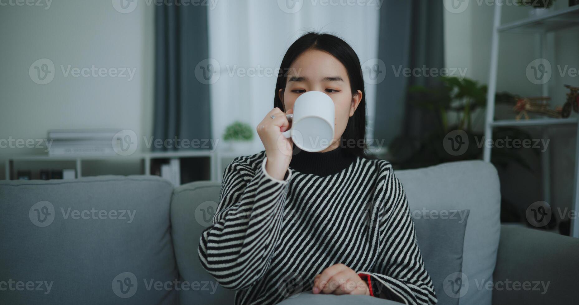 retrato de relajante joven mujer participación taza disfrutar oler café o té y Bebiendo con felicidad en Mañana mientras sentar en sofá en vivo habitación, gratis tiempo, toma descanso en casa, sonriendo foto