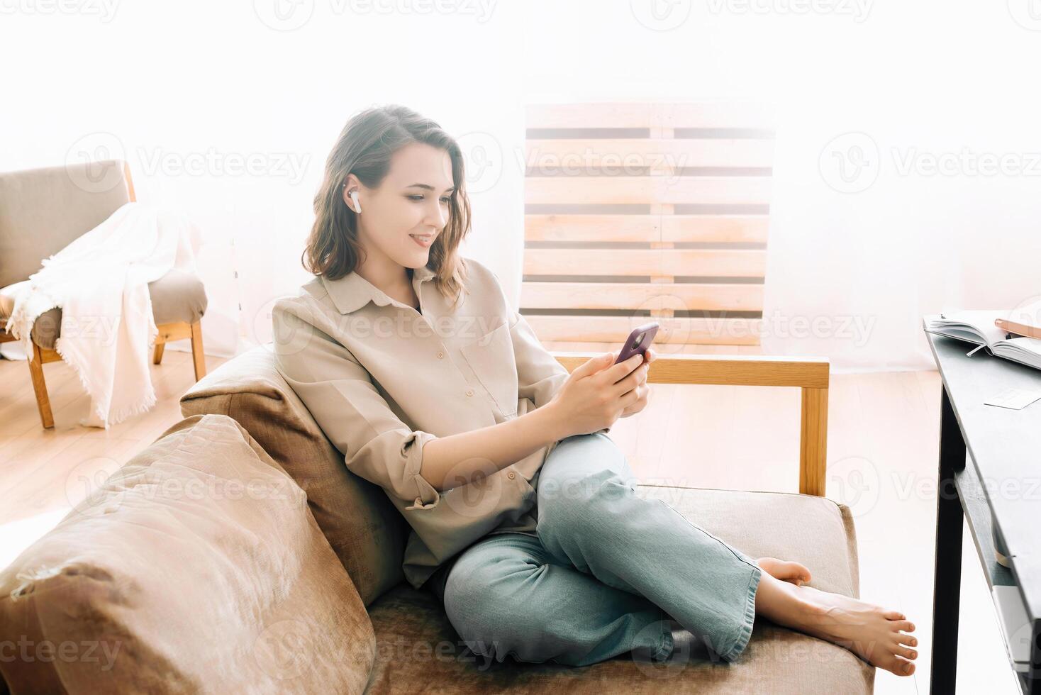 Glad millennial woman in wireless headphones looks at phone, reads messages, enjoys chatting. Communication, Digital Lifestyle, and Technology Happiness Concept. Wireless Connection Bliss. photo