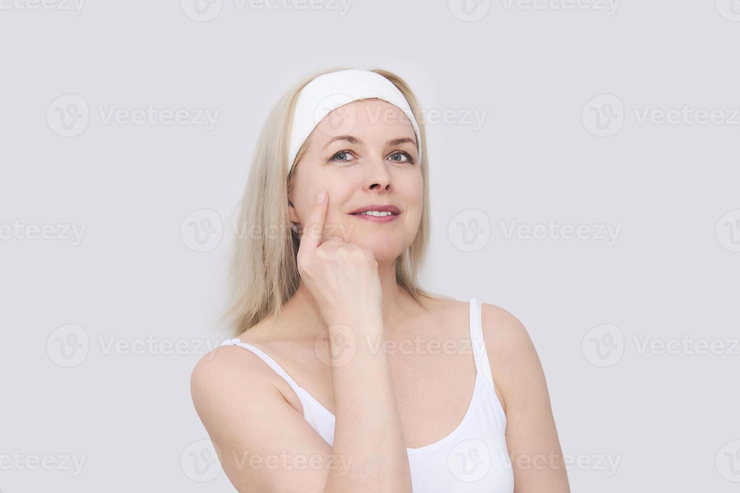 middle-aged woman in a hair band for beauty treatments points her finger at the condition of her facial skin, close-up portrait photo