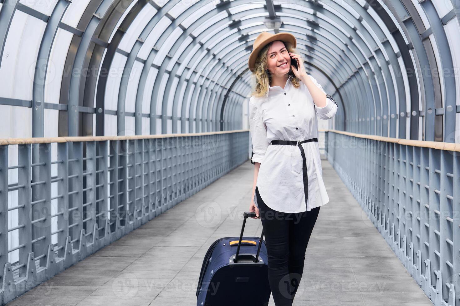 woman traveler with a suitcase smiling walks along the covered walkway gallery, speaks by phone photo