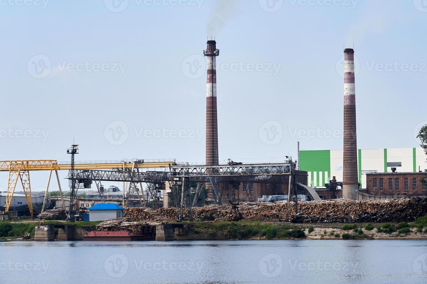 industrial paisaje, pulpa y papel molino con pilas de registros en el río banco foto