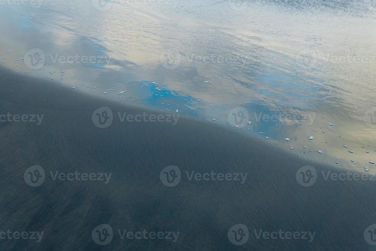 ocean beach with black volcanic sand, the sky is reflected in the rolled back wave of the surf photo