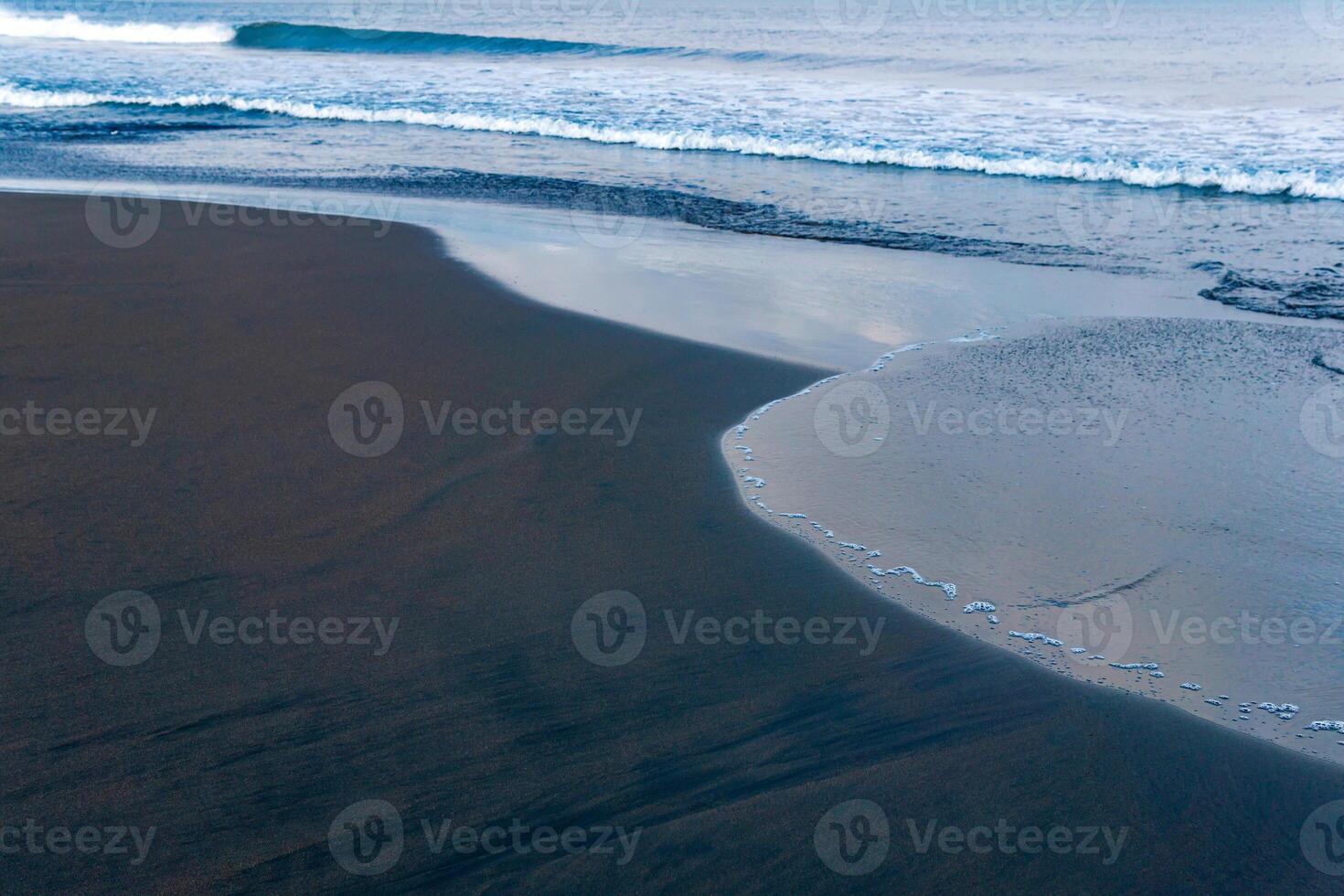 ocean beach with black volcanic sand, the sky is reflected in the rolled back wave of the surf photo