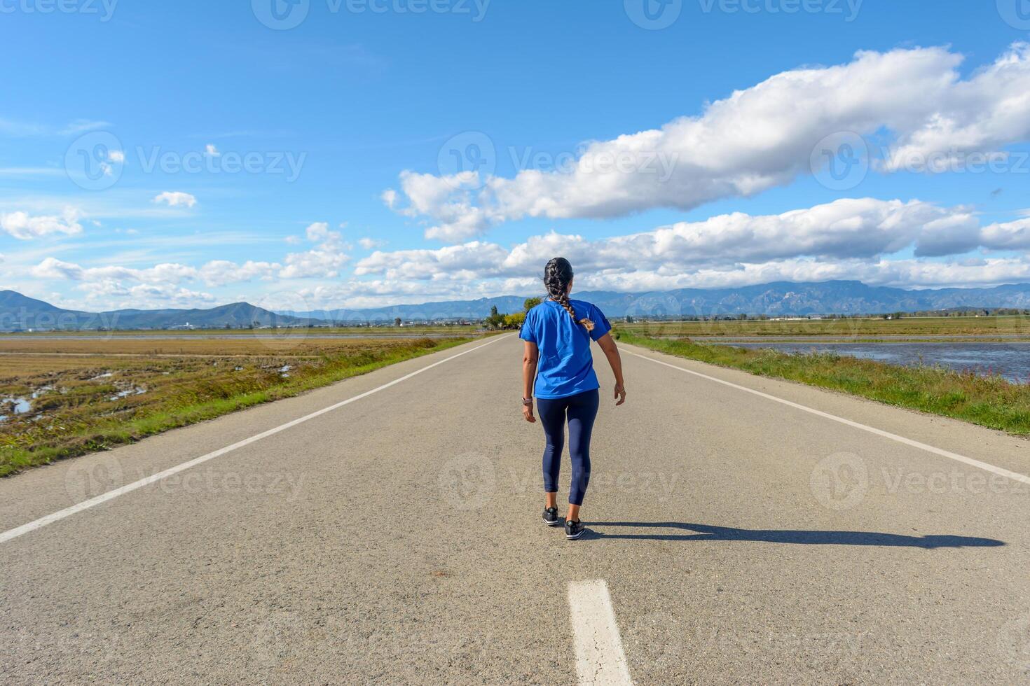 un persona camina abajo un Derecho la carretera con montañas y un claro cielo adelante, latina mujer caminando a lo largo solitario la carretera centrar en el ebro delta natural parque, tarragona, Cataluña, España foto