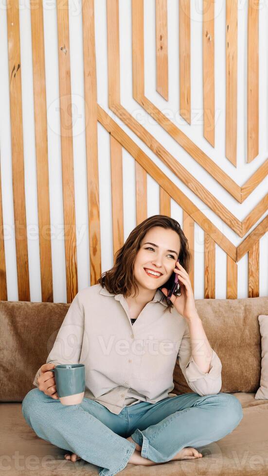 Cheerful Young Woman Talking on Phone, Relaxing on Sofa, Enjoying Refreshing Drink During Leisure Time. Cheerful young female calls by phone, speaks, sits on sofa, enjoys cup of drink in free time photo