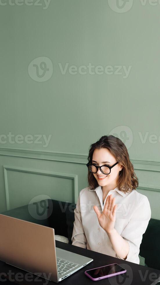 Cheerful Video Chat. Young Woman with Glasses Sharing Greetings. Expressive Online Interaction. Joyful Woman Waves in Laptop Call photo