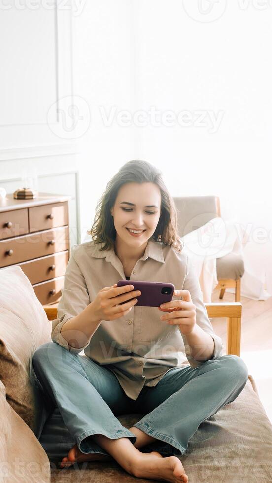 Joyful Millennial Leisure. Smiling young woman indulges in smartphone games, experiencing carefree enjoyment during her free time. Mobile Gaming and Relaxed Tech Fun. photo