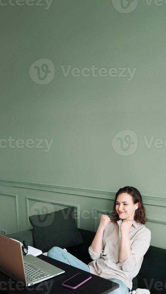 victorioso y celebrando. sonriente mujer gesticulando en un exitoso vídeo charla en su ordenador portátil. contento mujer en lentes es radiante como ella comunica en su computadora portátil, señalización victoria y éxito. foto