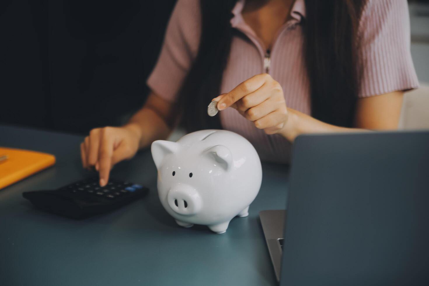 joven asiático mujer ahorro para Jubilación ahorro dinero mediante un cerdito banco y tomando notas en computadora portátil, ahorros concepto. foto