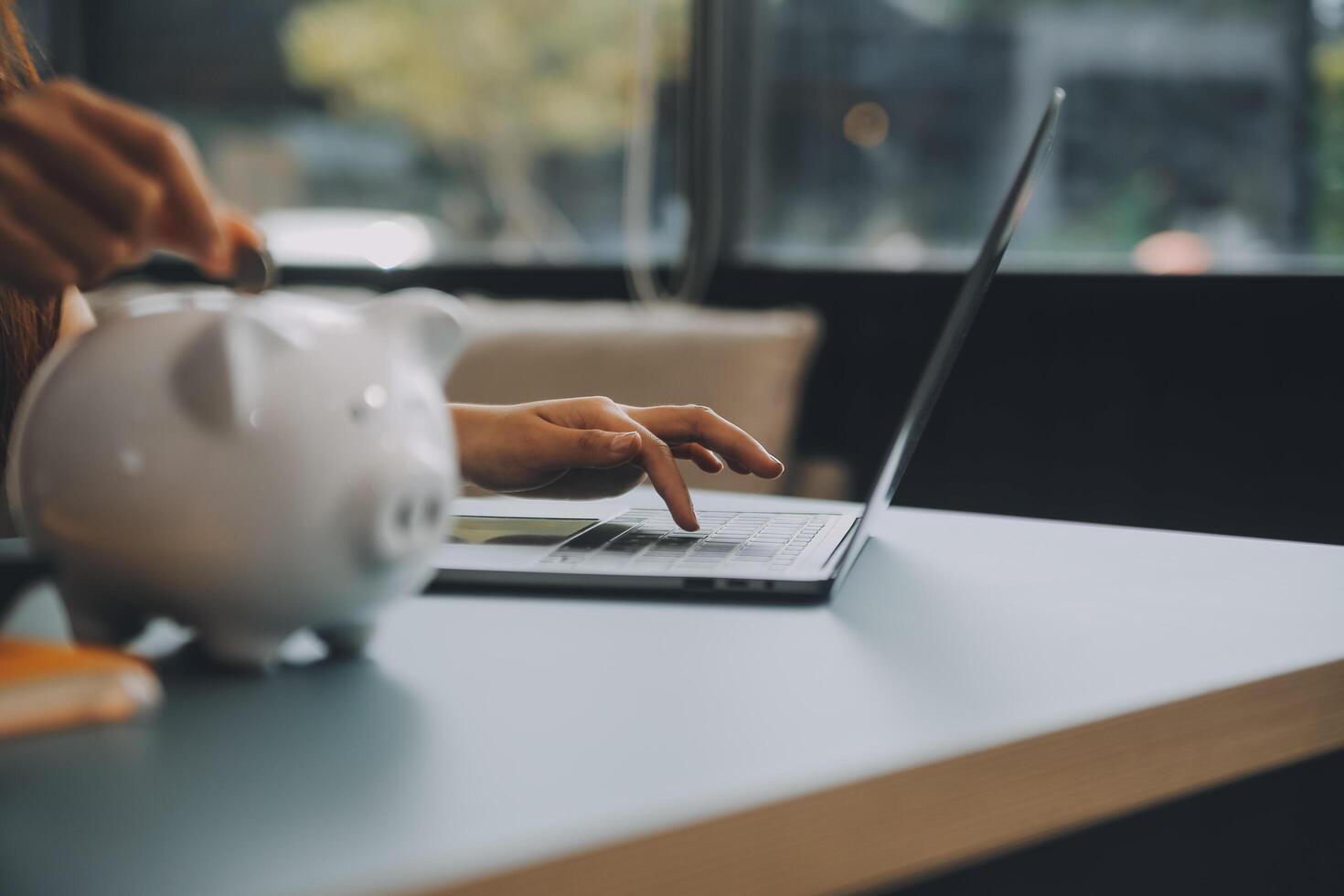 joven asiático mujer ahorro para Jubilación ahorro dinero mediante un cerdito banco y tomando notas en computadora portátil, ahorros concepto. foto