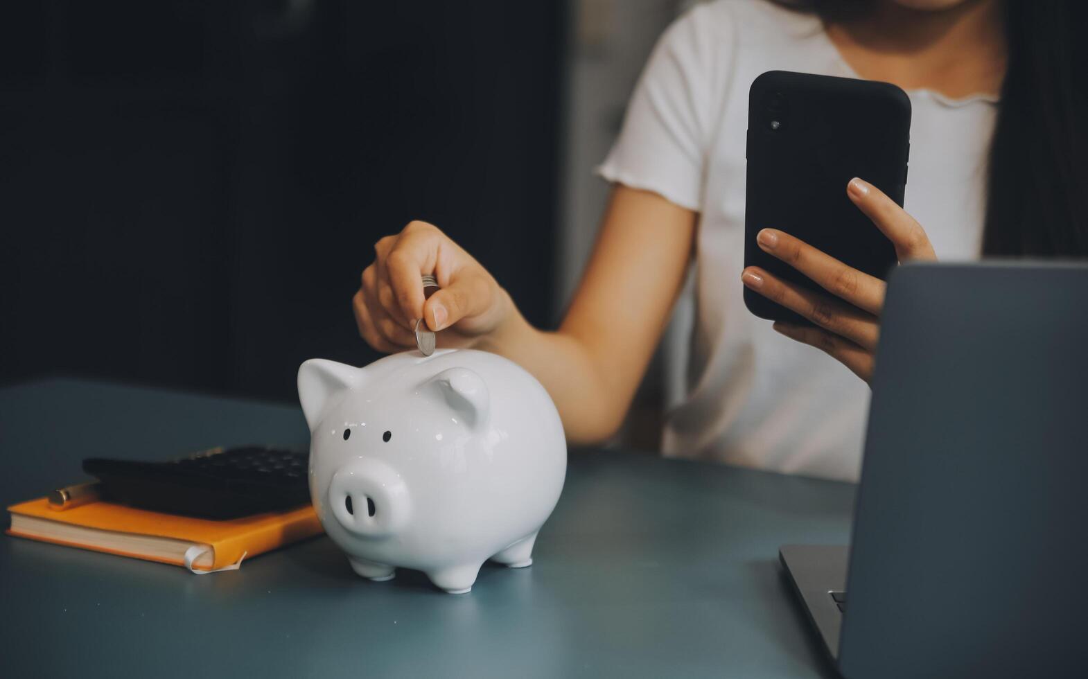 joven asiático mujer ahorro para Jubilación ahorro dinero mediante un cerdito banco y tomando notas en computadora portátil, ahorros concepto. foto