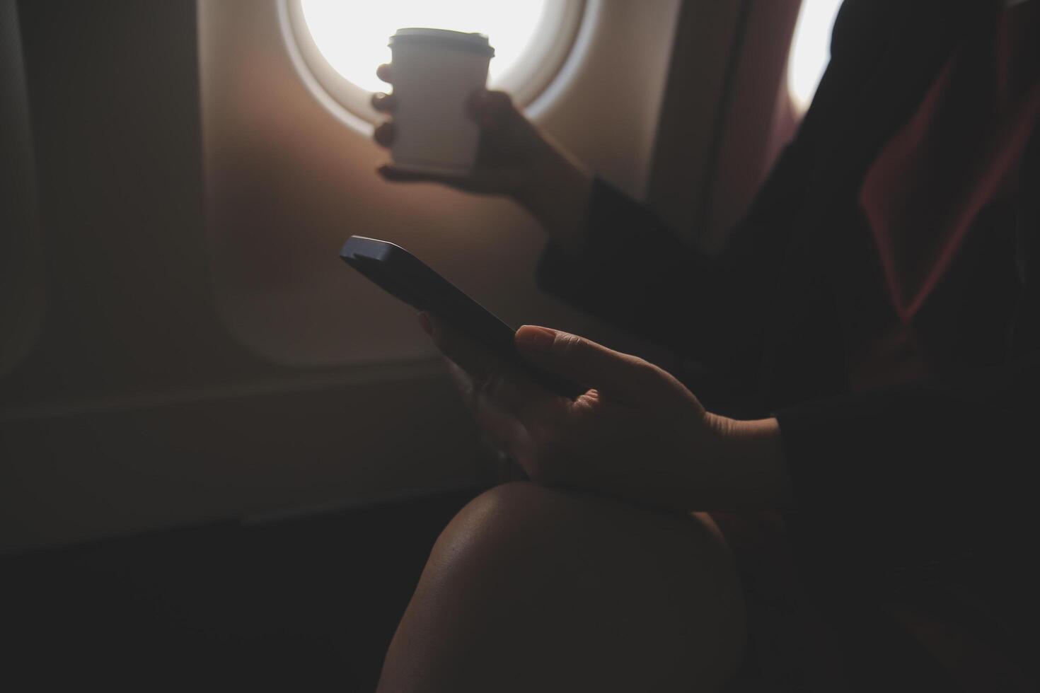 Blonde female tourist checking incoming notification on smartphone sitting on seat of airplane with netbook.Young businesswoman share media from telephone on laptop computer during plane flight photo