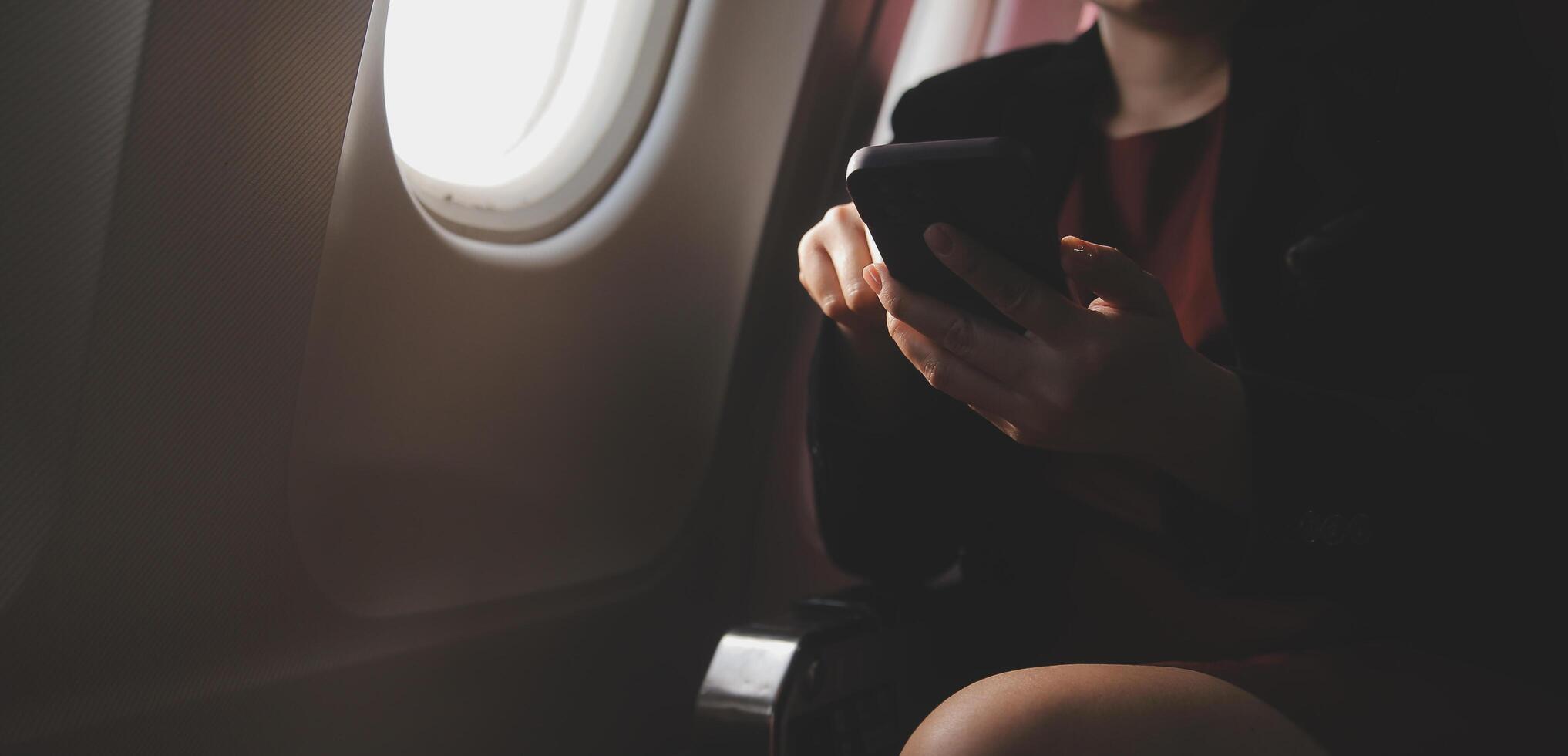 Blonde female tourist checking incoming notification on smartphone sitting on seat of airplane with netbook.Young businesswoman share media from telephone on laptop computer during plane flight photo