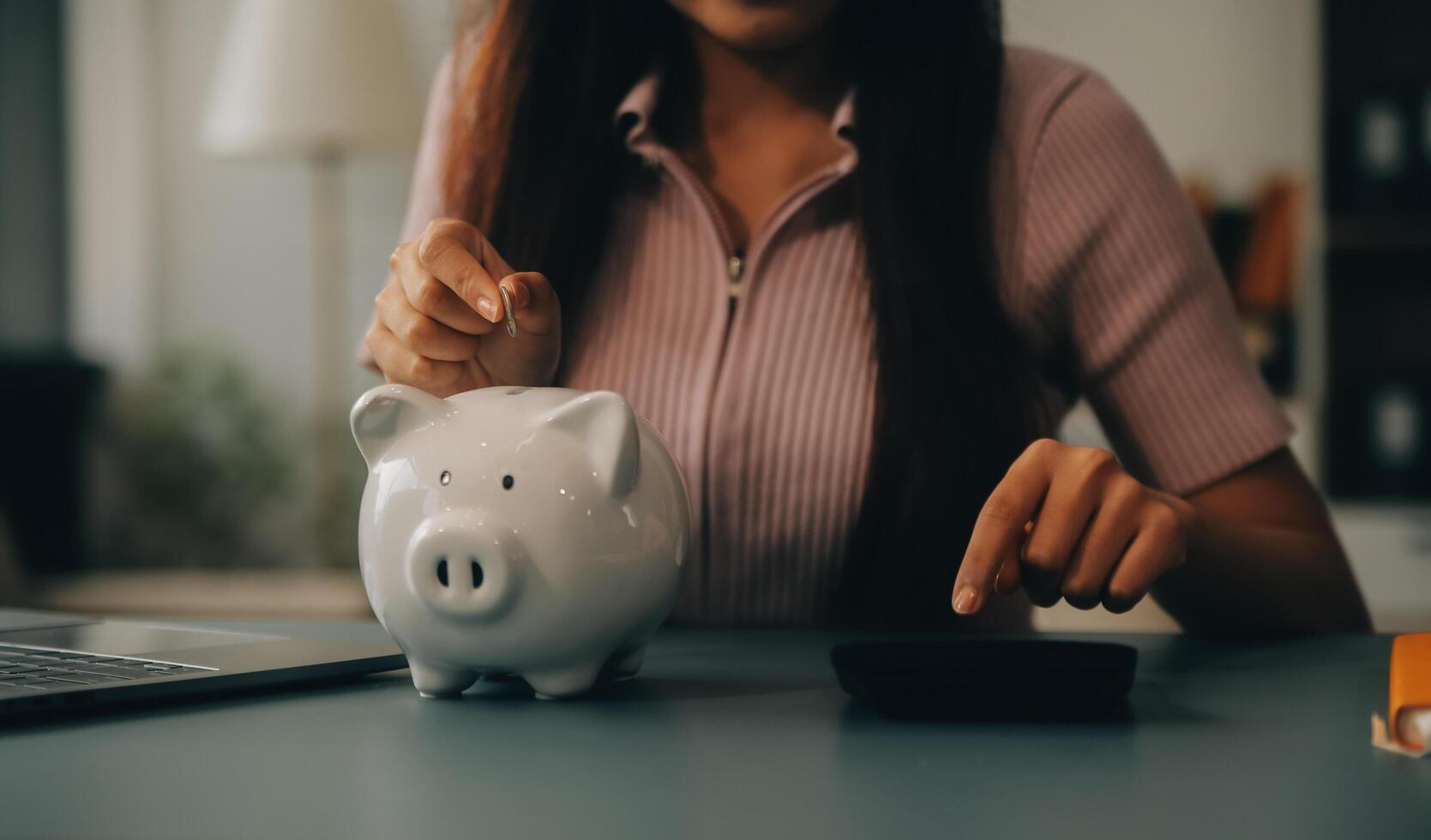 joven asiático mujer ahorro para Jubilación ahorro dinero mediante un cerdito banco y tomando notas en computadora portátil, ahorros concepto. foto