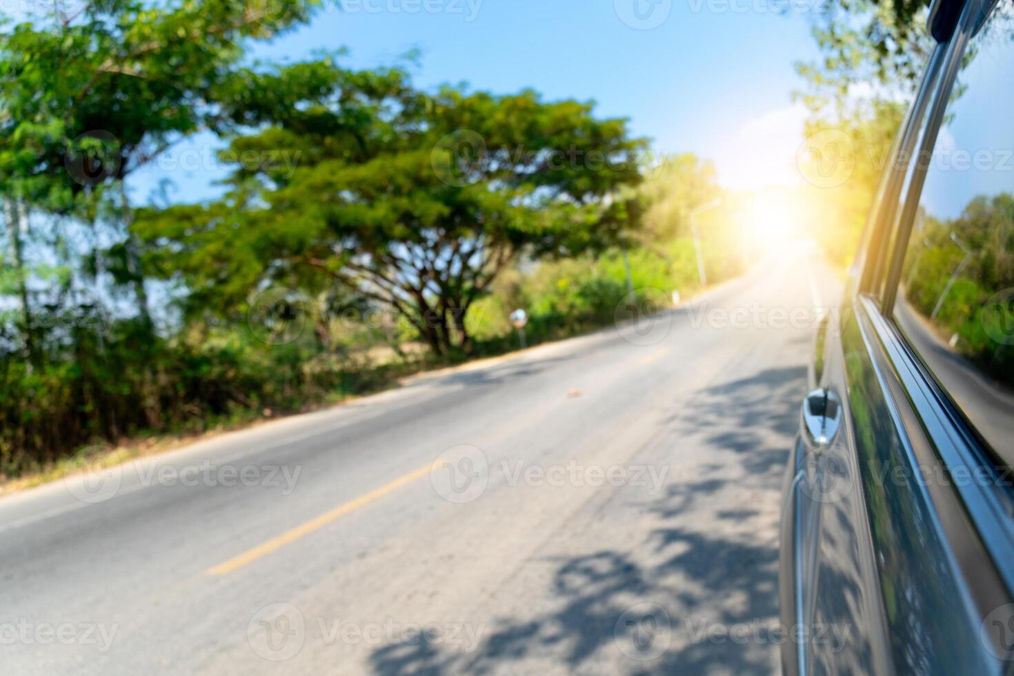 View on the side of car showing the windows and shiny light of car. Driving on asphalt road the side with nature of green trees. light coming straight from the destination. photo