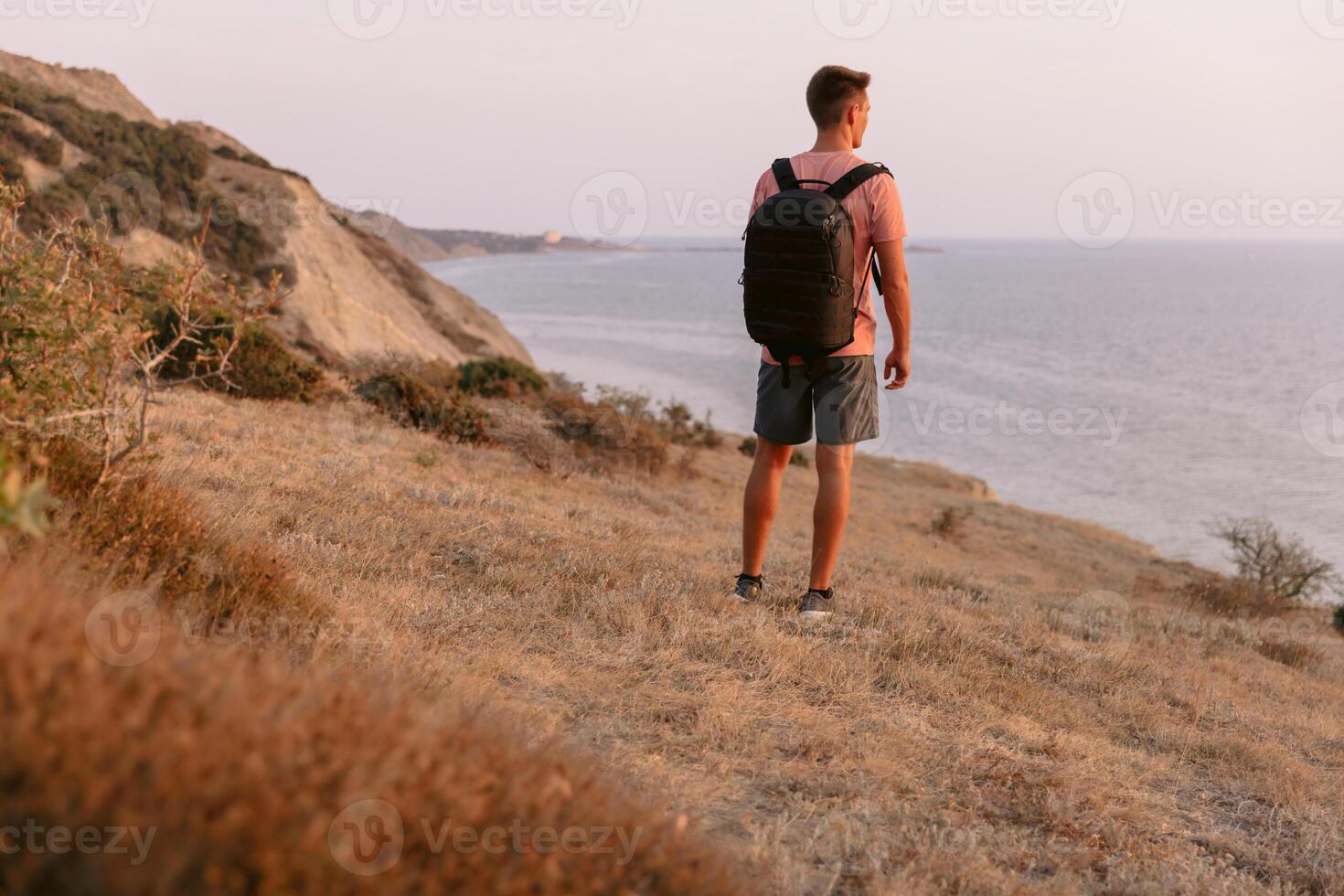 Man in a T-shirt and shorts with backpack walking on the coastline with warm sunset light. photo