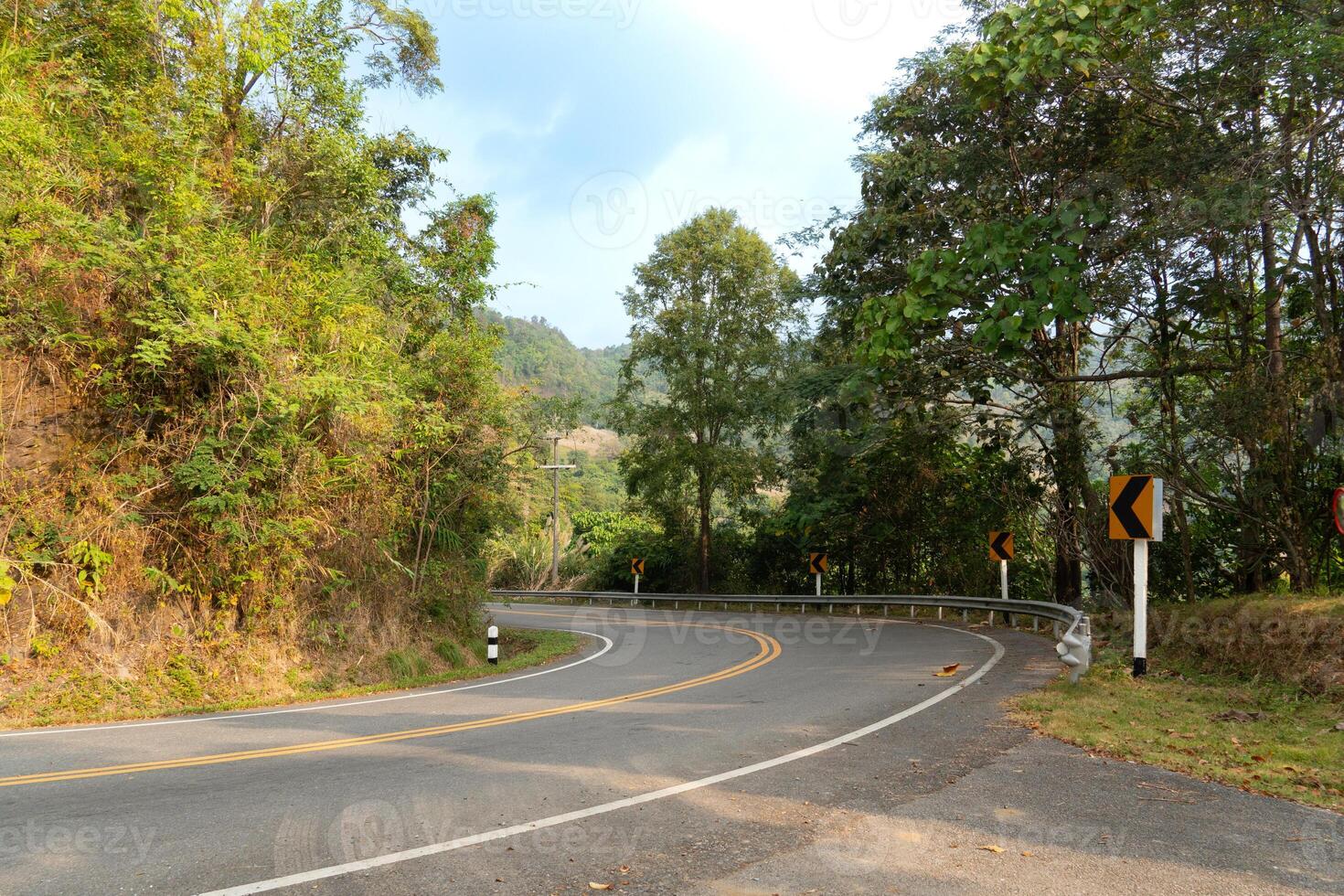 Curved downhill asphalt road with mountains next to it and spring foliage. With sign board for control curved. Forest trees under the blue sky. photo
