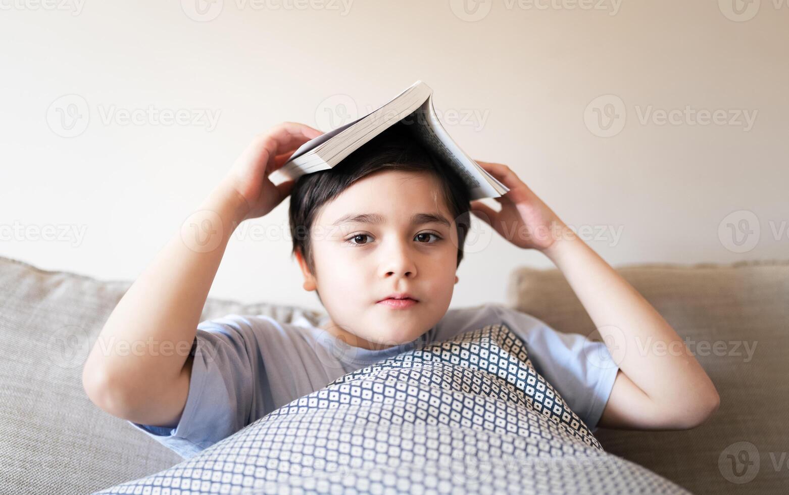 alegre colegio niño chico participación libro en su cabeza sentado en sofá en vivo habitación, atrás a colegio concepto joven niño leyendo libro o haciendo hogar trabajo a hogar en fin de semana, un chico sonriente y mirando a cámara foto