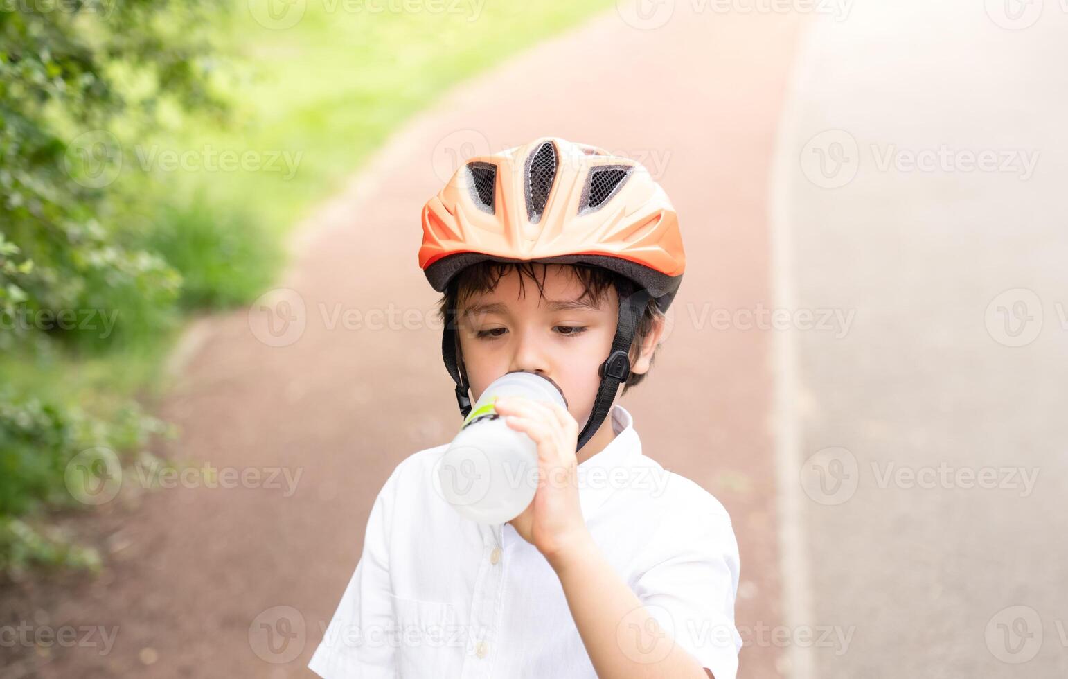 Healthy child boy wearing a bike helmet and drinking water,  kid Thirsty boy holding a bottle of water wearing a cycling helmet riding a bicycle in the park, Concept for safety, childhood health care photo