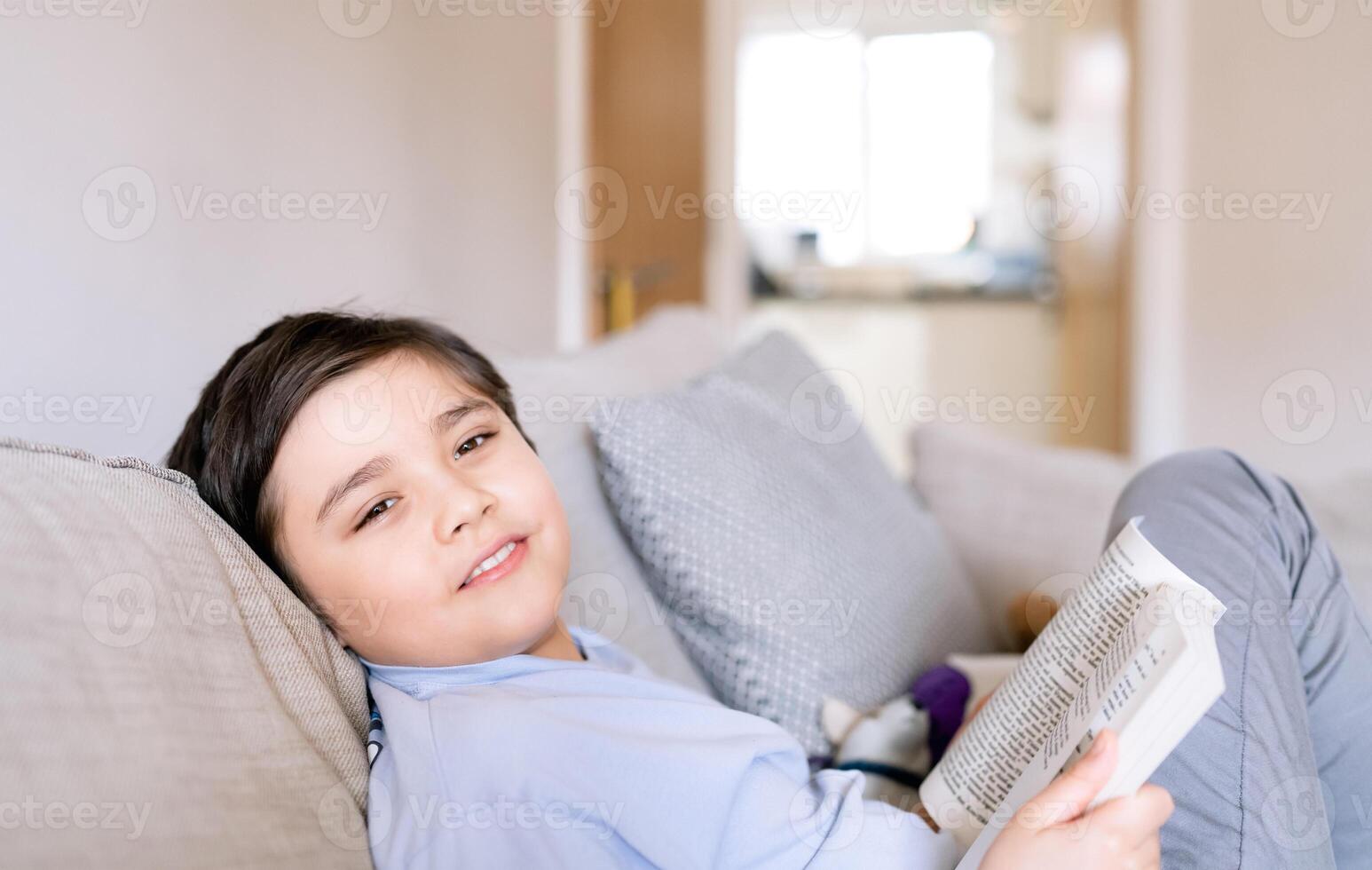 alegre colegio niño chico participación libro sentado en sofá en vivo habitación, atrás a colegio concepto joven niño leyendo libro o haciendo hogar trabajo a hogar en fin de semana, un chico sonriente y mirando a cámara foto