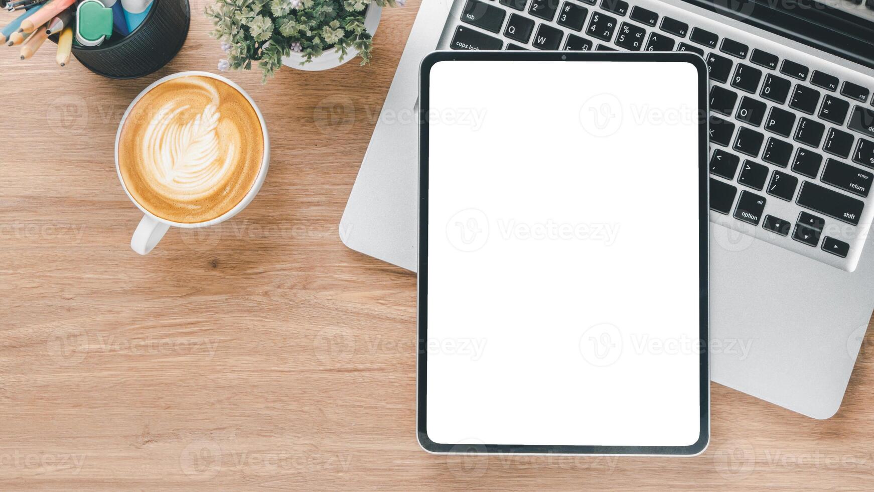 An organized work desk featuring a laptop, digital tablet with a blank screen, a cup of cappuccino, and office supplies from an overhead view. photo