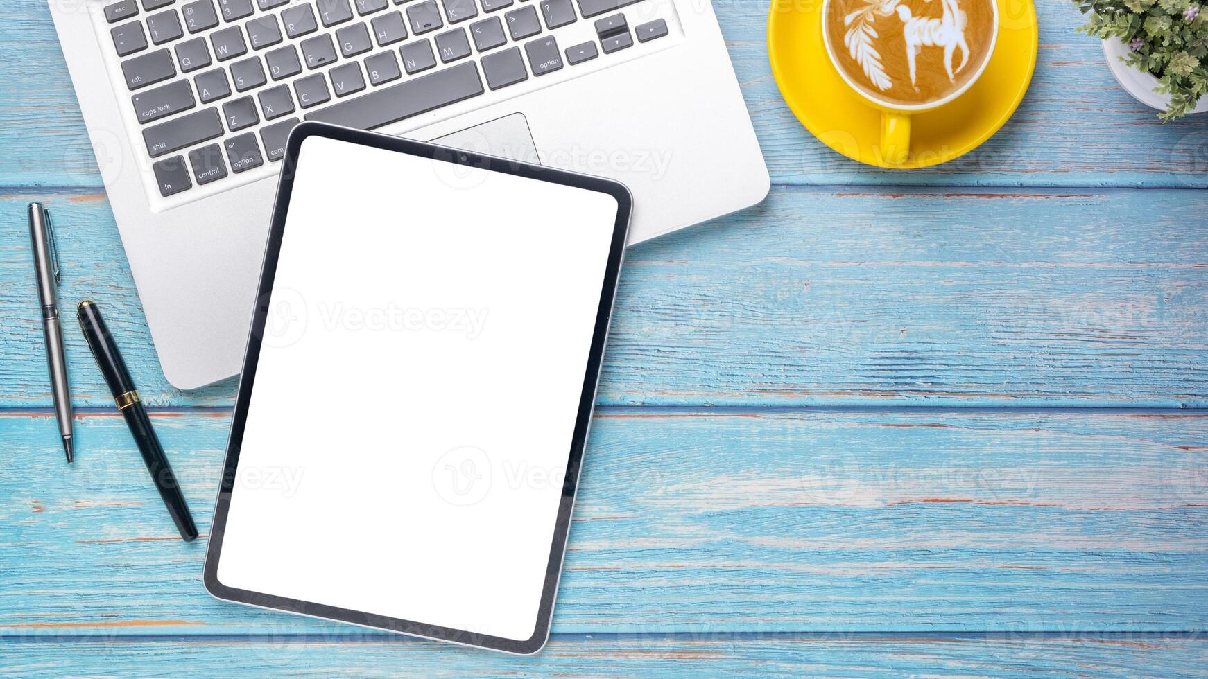 A clean and organized mobile work setup featuring a tablet with a blank screen next to a laptop, pen, and a decorative cappuccino on a blue wooden table. photo