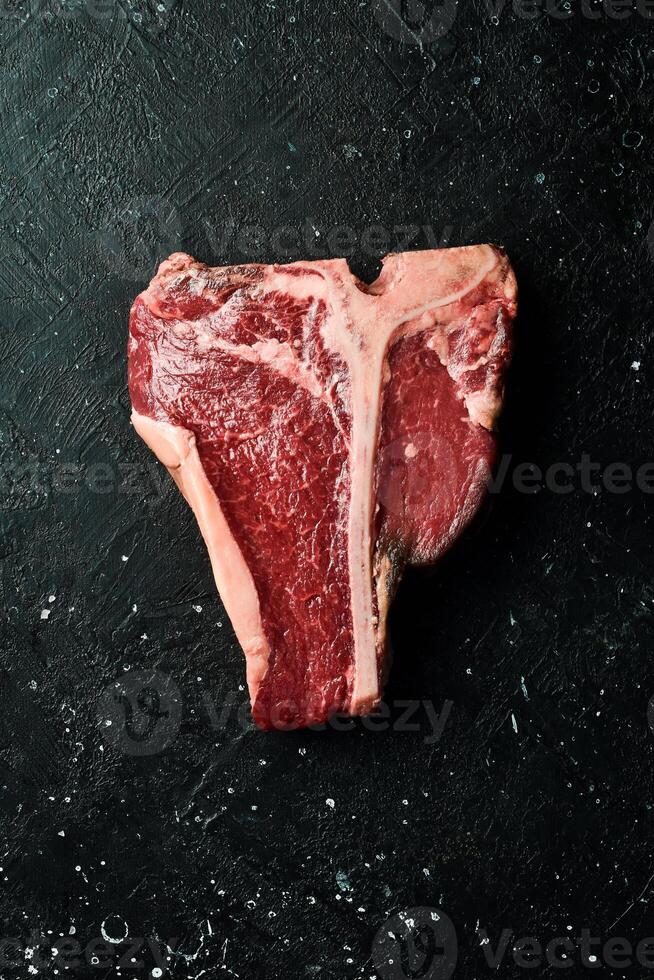 Large raw t-bone steak on a wooden board with seasonings and pepper. On a black stone table. Top view. photo