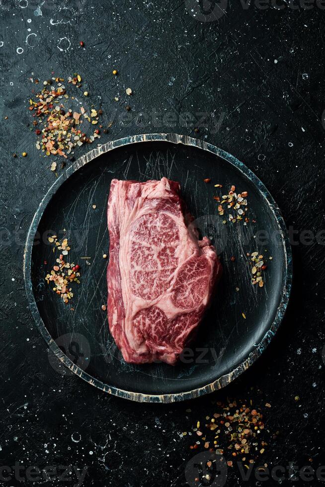 Raw Steak Ribeye Black Angus on a wooden plate ready to be cooked. On a black stone background. Rustic style. photo